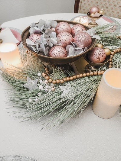 Festive table centerpiece with pink and gold ornaments, pine branches, a string of gold beads, and lit candles.