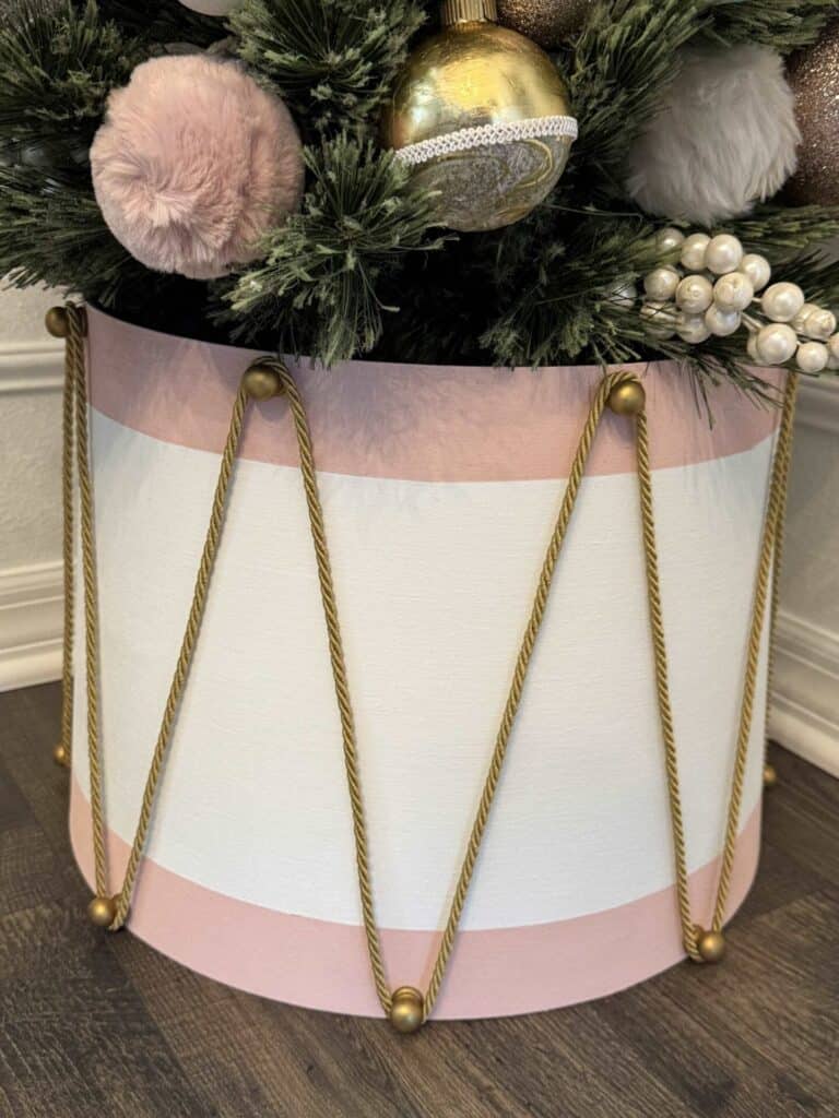 A decorative drum-shaped planter with pink and white stripes, filled with artificial greenery and embellished with ornaments, sits on a wooden floor.