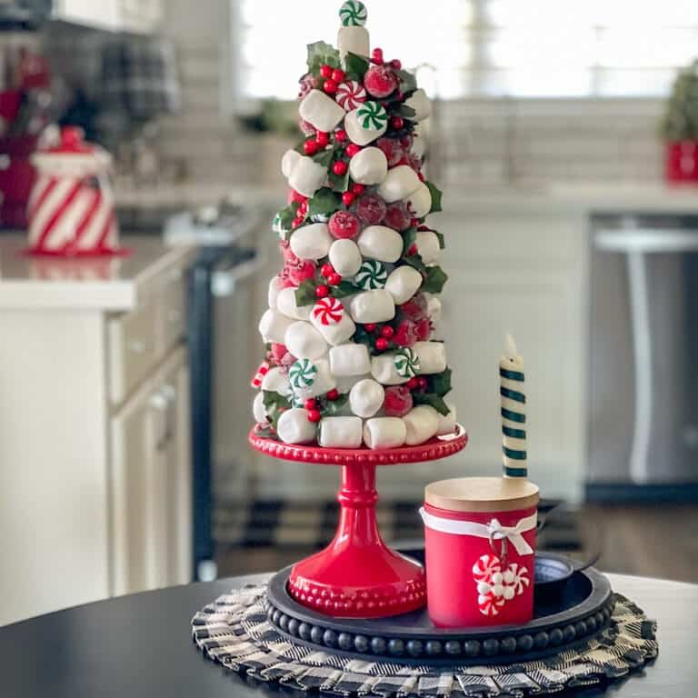 Festive candy tree on a red stand, adorned with marshmallows, peppermints, and small red berries, placed on a black tray with a red candle in a kitchen setting.