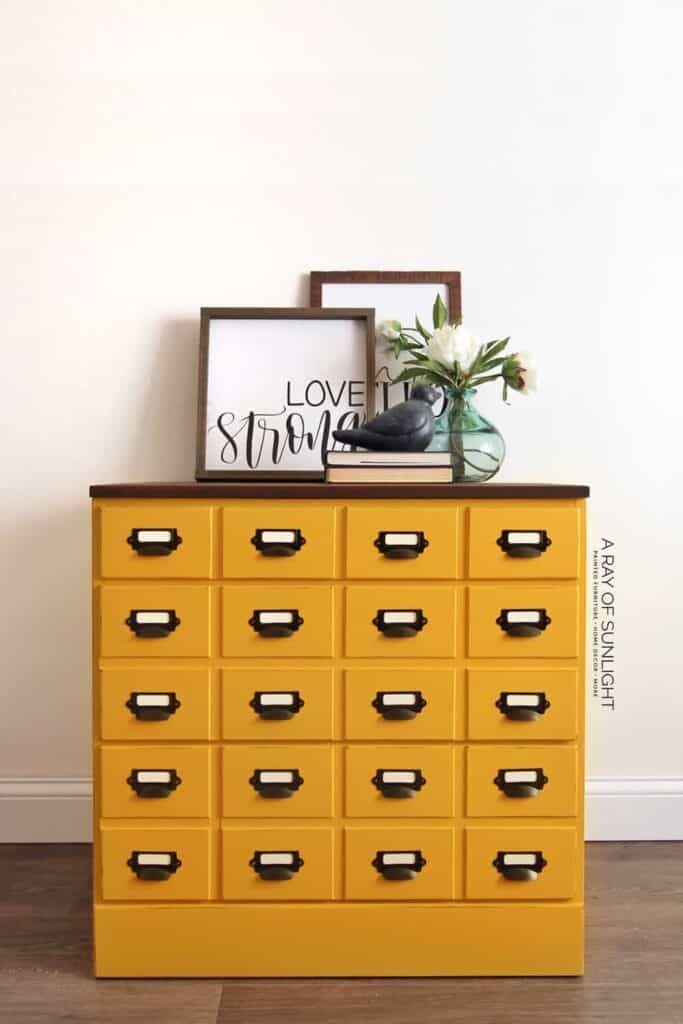 Yellow dresser with multiple drawers, decorated with framed art and flowers in a vase on top.