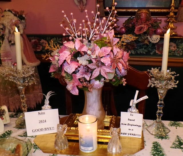 A festive table display features a pink floral centerpiece, candles, angel figurines, and signs saying "Kiss 2024 GOOD BYE!!" and "To Sweet New Beginnings in A NEW YEAR!!!!.