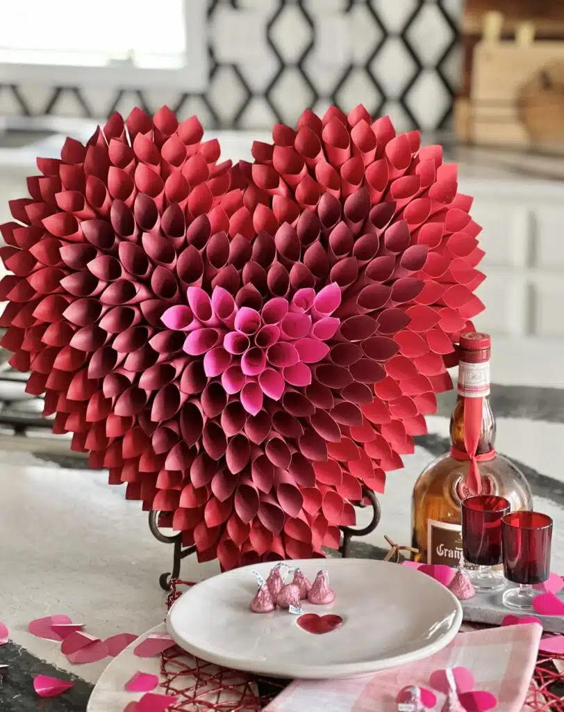 Red and pink paper heart-shaped decoration on a table with a bottle, two glasses, rose petals, and a plate with chocolates. Kitchen backsplash visible in the background.