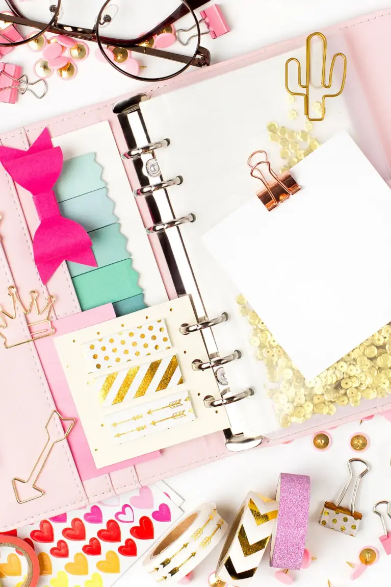 An open planner with decorative stickers, sticky notes, paper clips, and glasses on a white surface.