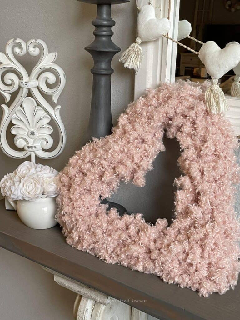 Pink fluffy heart-shaped wreath on a shelf with a white vase of roses, decorative elements, and a string of heart ornaments.