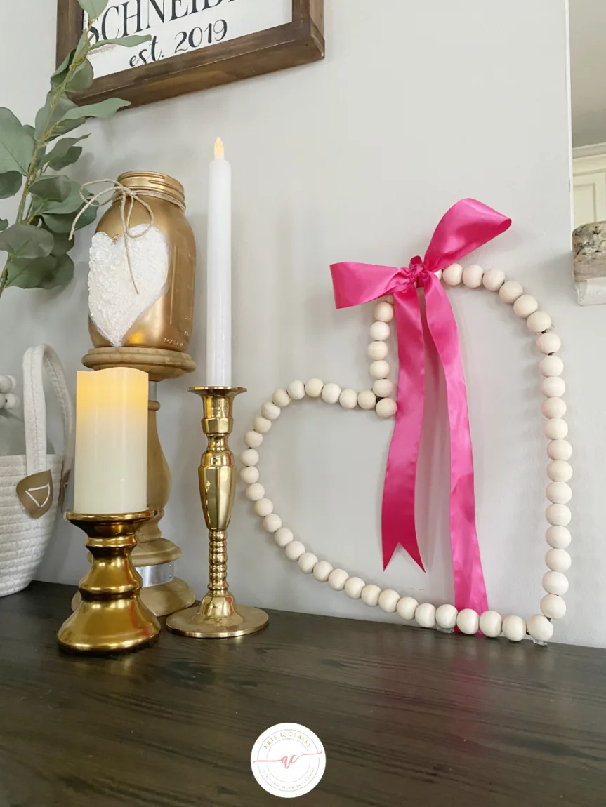 Candles on brass holders, a white beaded heart with a pink ribbon, and decorative jars sit on a dark wooden surface. A framed sign and greenery are in the background.