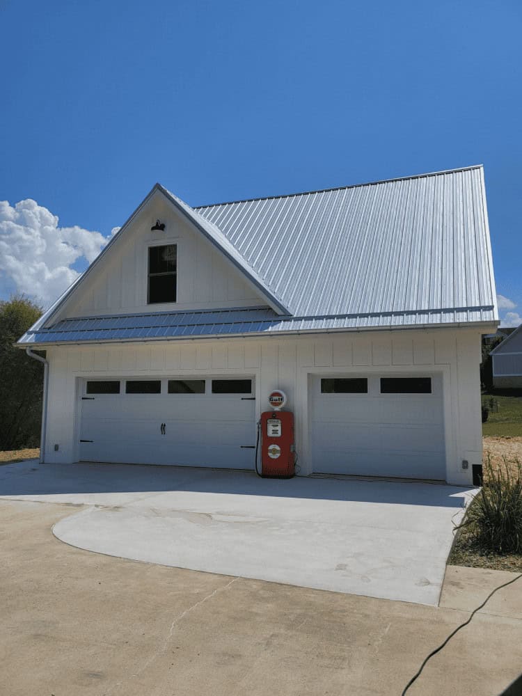 White garage with a metal roof and two garage doors. A vintage red gas pump is placed between the doors. Blue sky and a few clouds in the background.