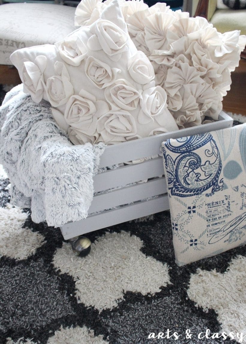 White crate with floral-patterned pillows and soft blanket on a fluffy rug.