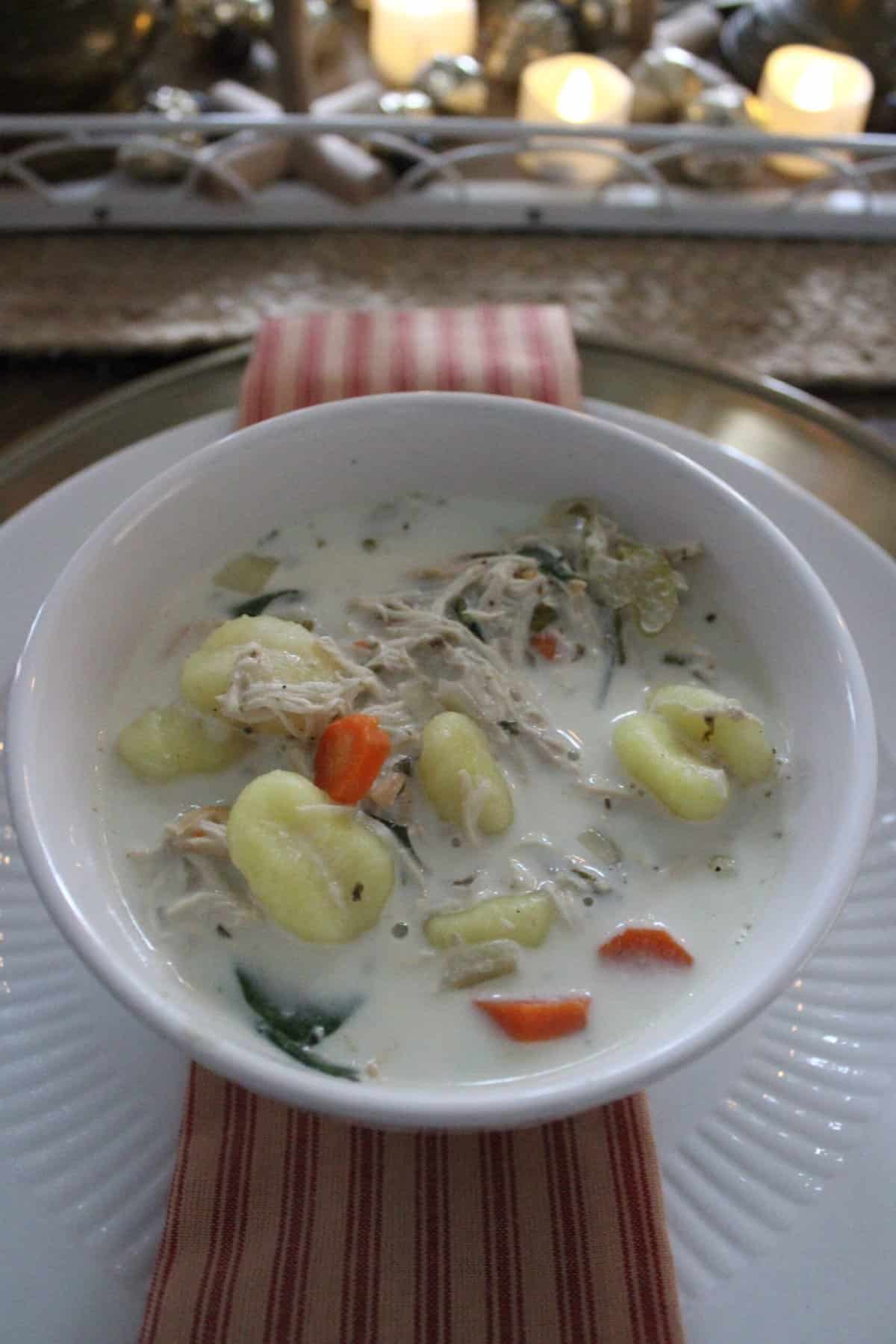 A bowl of creamy chicken soup with gnocchi, carrots, celery, and herbs, placed on a red-striped cloth. Candles and decorations are softly blurred in the background.