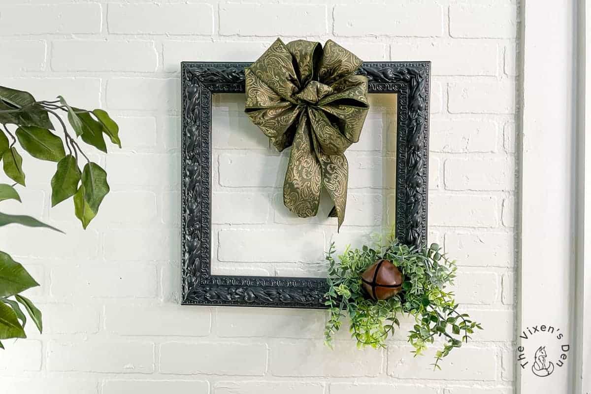 A decorative black frame with an ornate green bow and greenery, featuring a small brown bell, hangs on a white brick wall next to a plant.