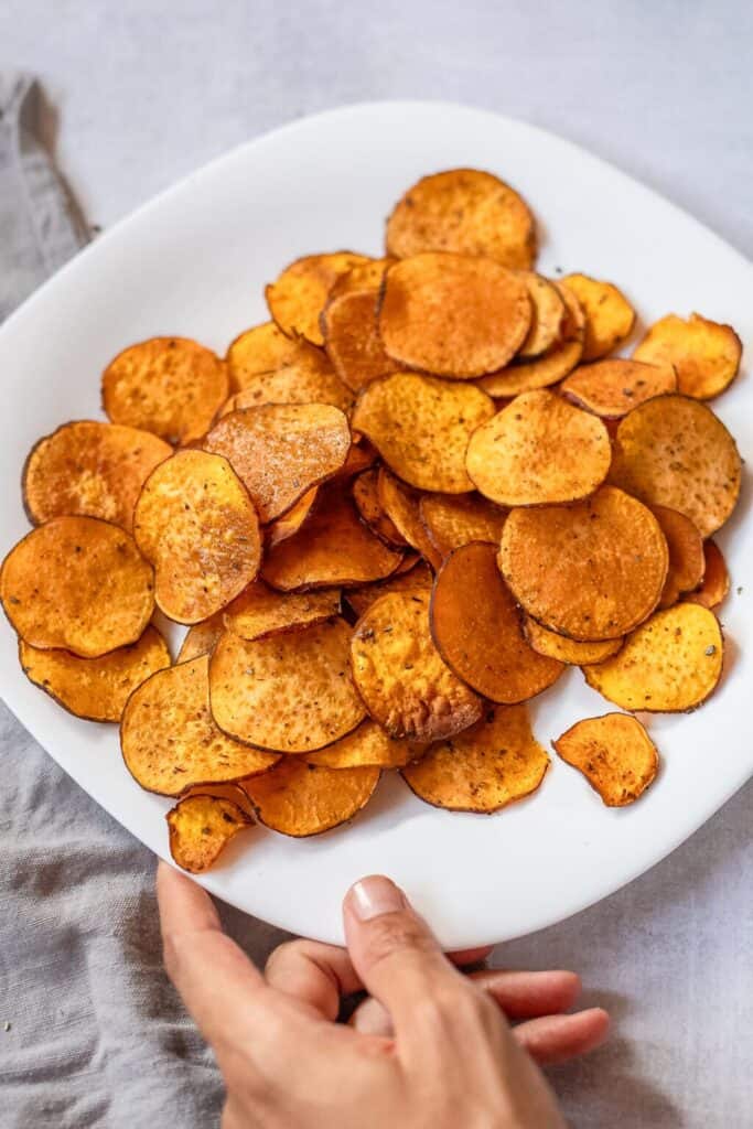 A hand holding a white plate with a pile of crispy, round sweet potato chips on a light-colored surface.