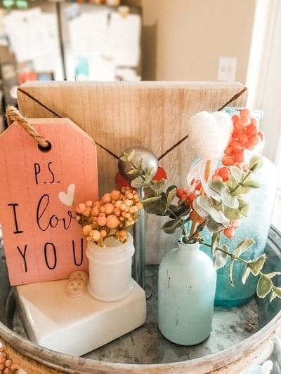 A decorative display features a pink sign reading "P.S. I love you," two vases with flowers and leaves, and a wooden board on a metal tray.