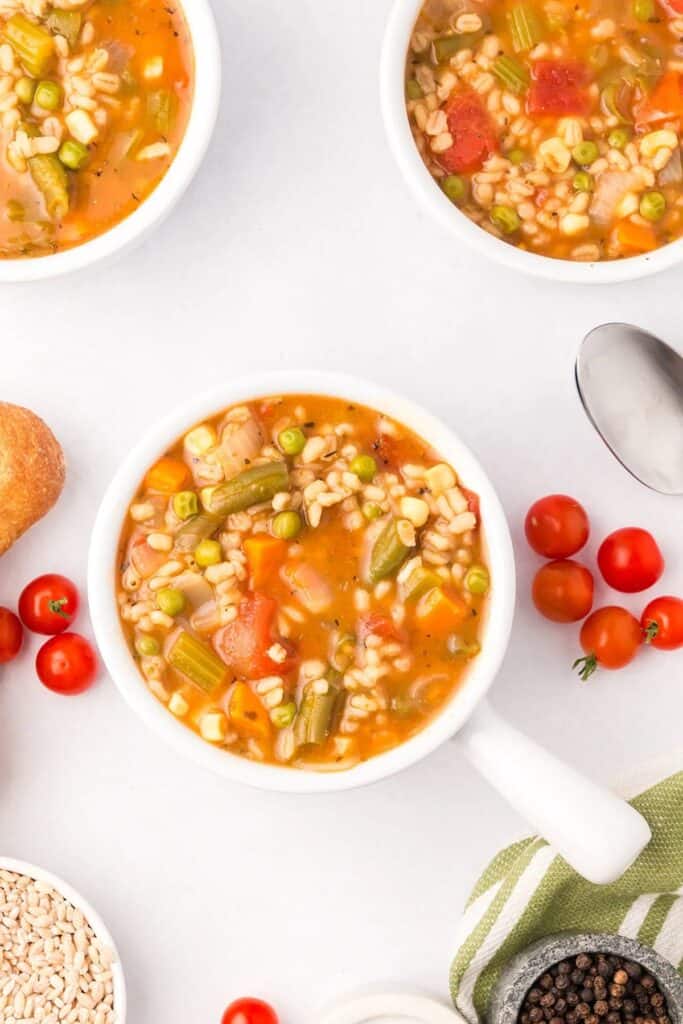 Bowl of vegetable and barley soup surrounded by cherry tomatoes, a bread roll, a spoon, and a bowl of peppercorns on a white surface.