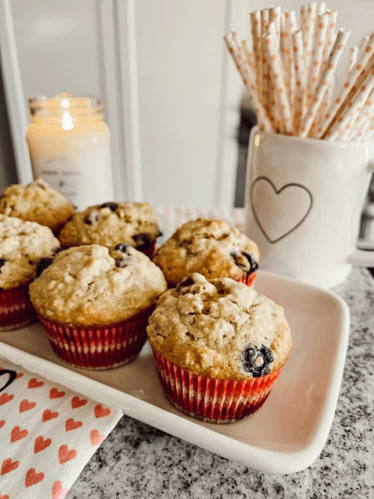 Blueberry muffins on a white plate with a lit candle, a mug containing straws, and a heart-patterned napkin on a grey countertop.