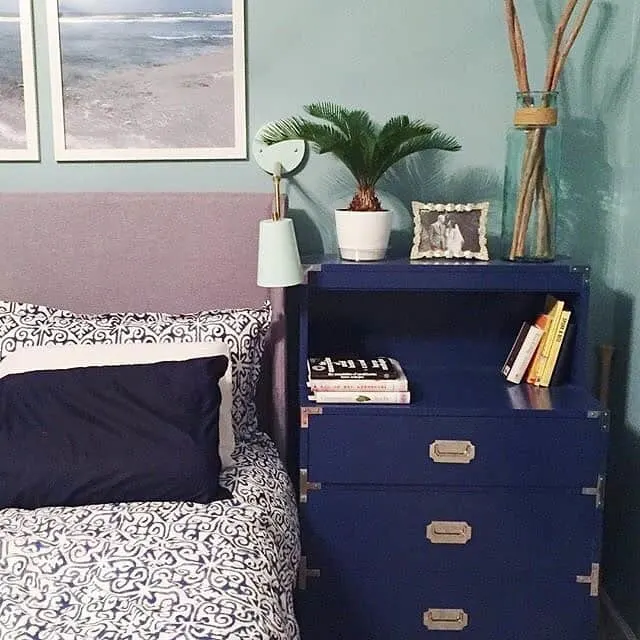 Bedroom corner with a blue dresser, books, and a potted plant. A bed with patterned sheets and a navy pillow is beside it. Artwork and a framed photo are on the wall.