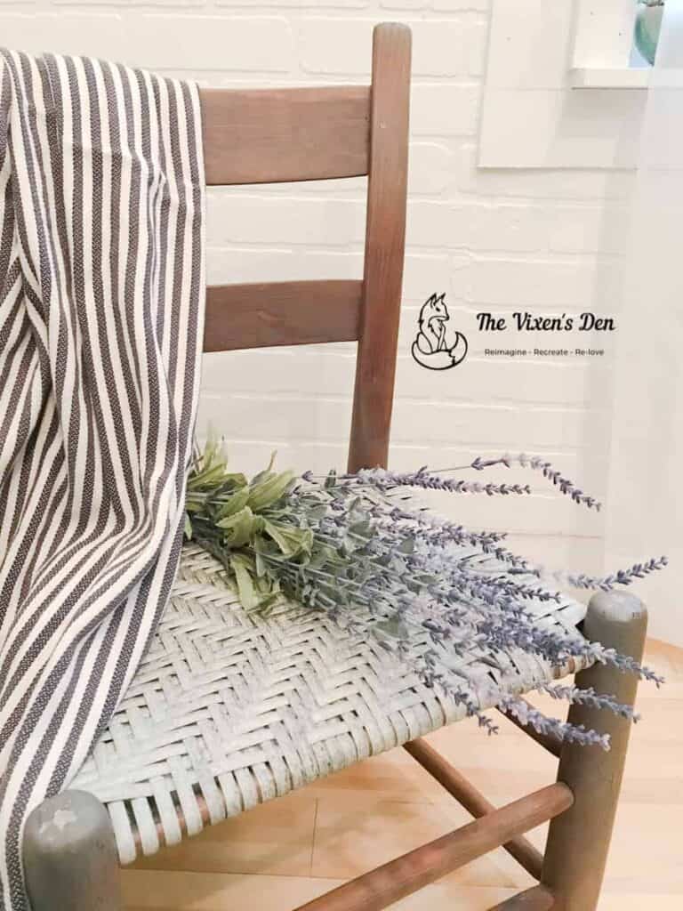 Wooden chair with a woven seat, decorated with a striped cloth and a bundle of lavender, placed against a white brick wall with a logo that reads "The Vixen's Den.