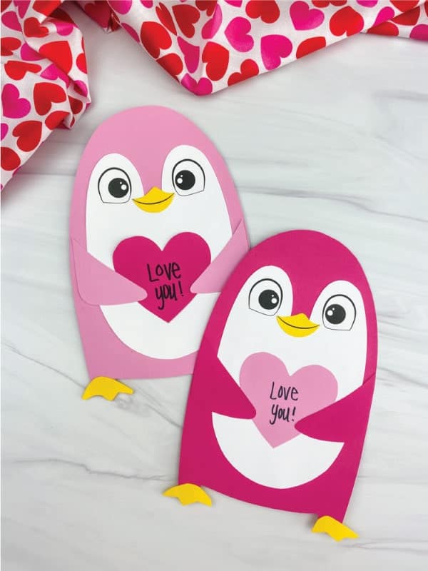 Two paper penguin crafts holding heart shapes with "Love you!" written on them, placed on a light surface with red and pink heart-patterned fabric in the background.