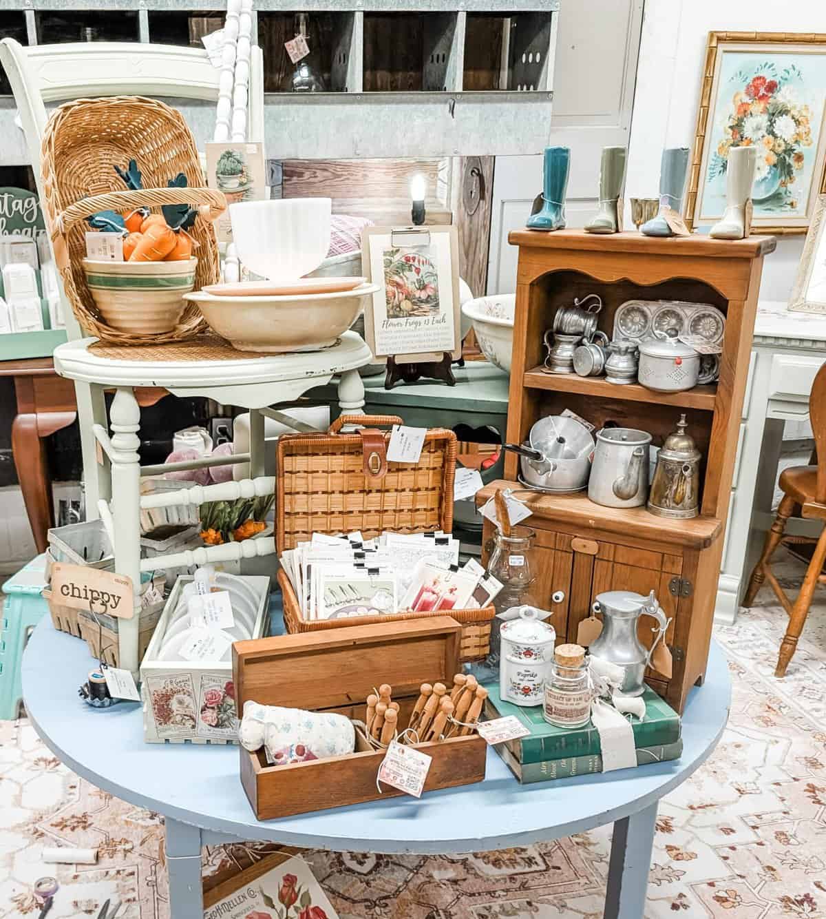 A vintage shop display featuring baskets, wooden cabinets, ceramics, books, and various kitchen items arranged on tables in a decorative manner.