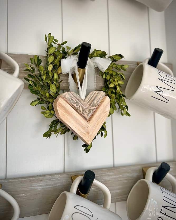 Wooden heart with ribbon hangs on green wreath among white mugs on hooks against a white wall.