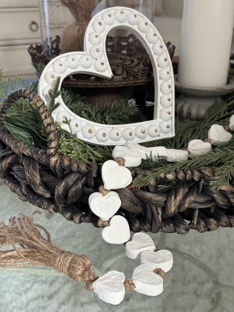 A wicker tray holds greenery, a white decorative heart, and a string of small white heart shapes with twine, placed on a glass surface.