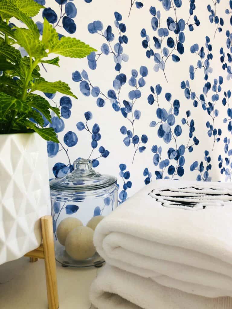 A plant in a geometric pot, a glass jar with two balls, and folded white towels are placed against a blue and white floral wallpaper.