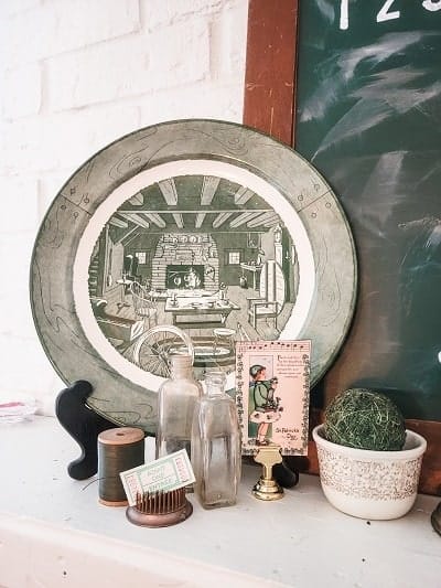 A decorative plate with a rustic scene sits on a shelf, accompanied by vintage bottles, a spool of thread, a small plant in a bowl, and a candle holder with a card.