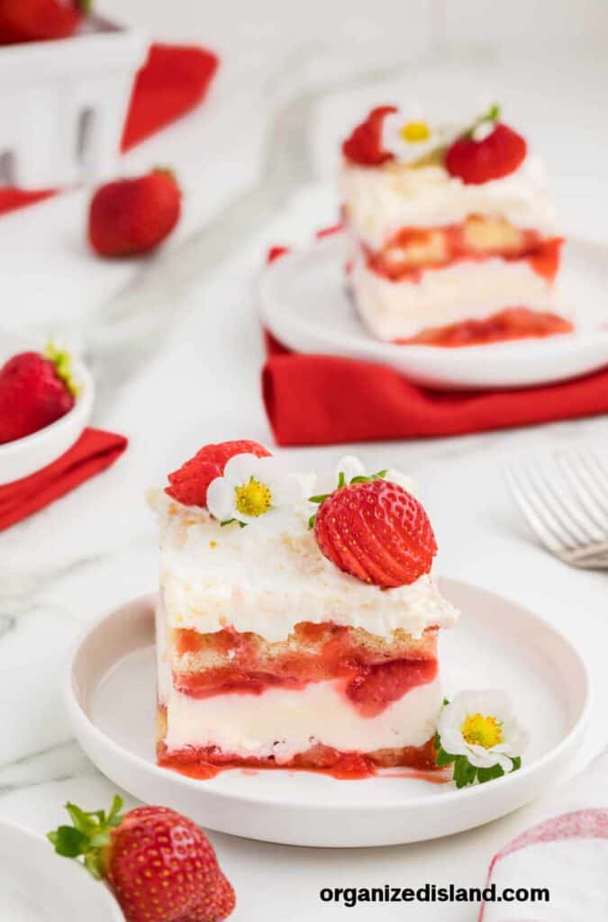 A slice of strawberry shortcake on a white plate, topped with whipped cream, strawberries, and a small white flower. Additional strawberries are scattered around on a white surface.