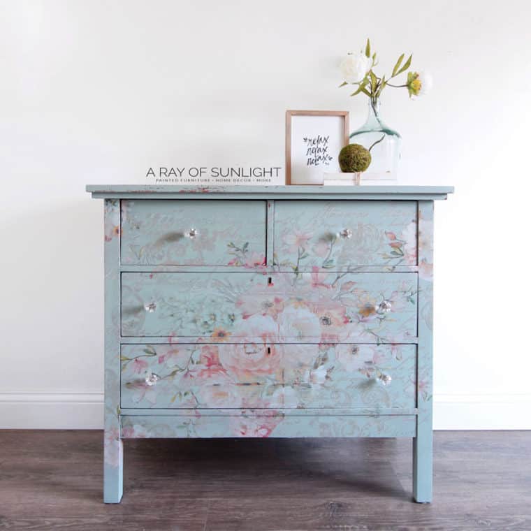 A blue floral-patterned dresser with four drawers, decorative items on top including a vase with flowers, and a framed print.