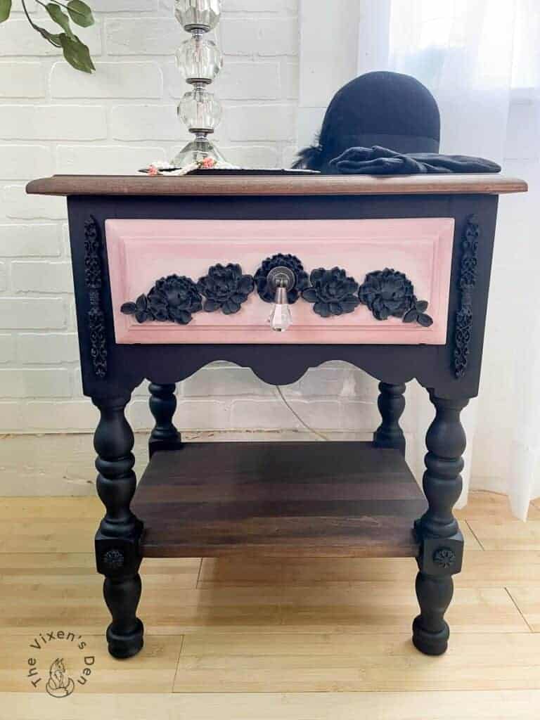 Wooden side table with a light pink drawer featuring black floral decorations and a crystal knob. A lamp and black hat rest on top. Brick wall and sheer curtain background.