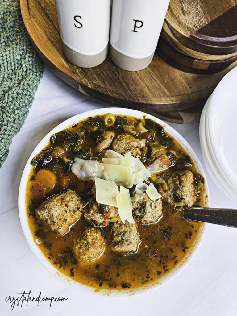 A bowl of soup with meatballs, greens, and shaved cheese. A spoon rests in the bowl. Salt and pepper shakers and a stack of plates are in the background.