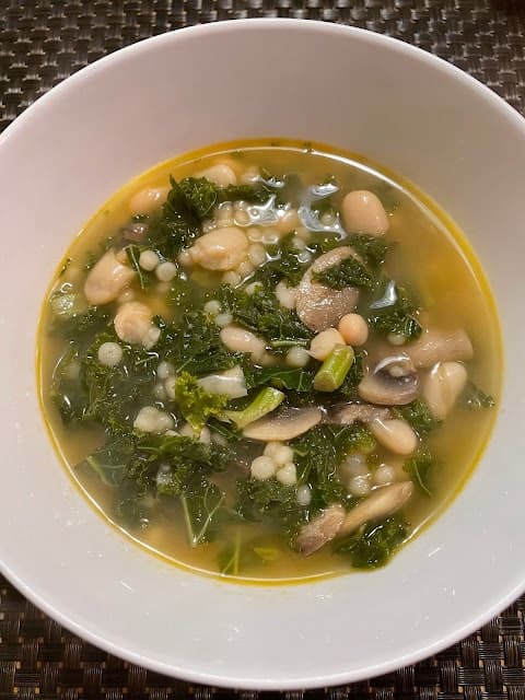 Bowl of soup with white beans, kale, mushrooms, and small pasta in broth.