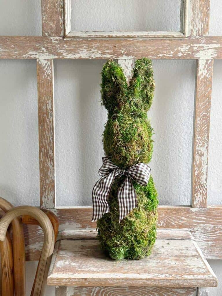 A moss-covered bunny-shaped figure with a plaid ribbon sits on a wooden shelf against a rustic window frame backdrop.