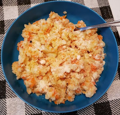 A blue bowl filled with mashed root vegetables, topped with grated cheese, on a black and white checkered tablecloth. A metal spoon is placed on the side of the bowl.