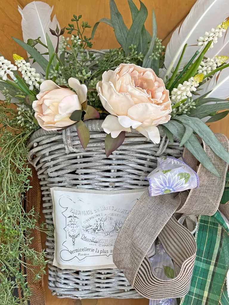 A wicker basket with light pink flowers, greenery, and feathers is adorned with ribbon and fabric. A decorative label is attached to the basket.