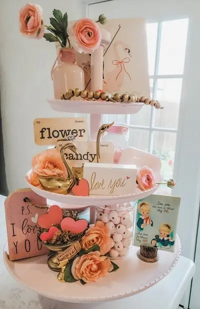 A two-tiered tray with pink and orange roses, decorative signs, a swan figurine, small jars, and candies.