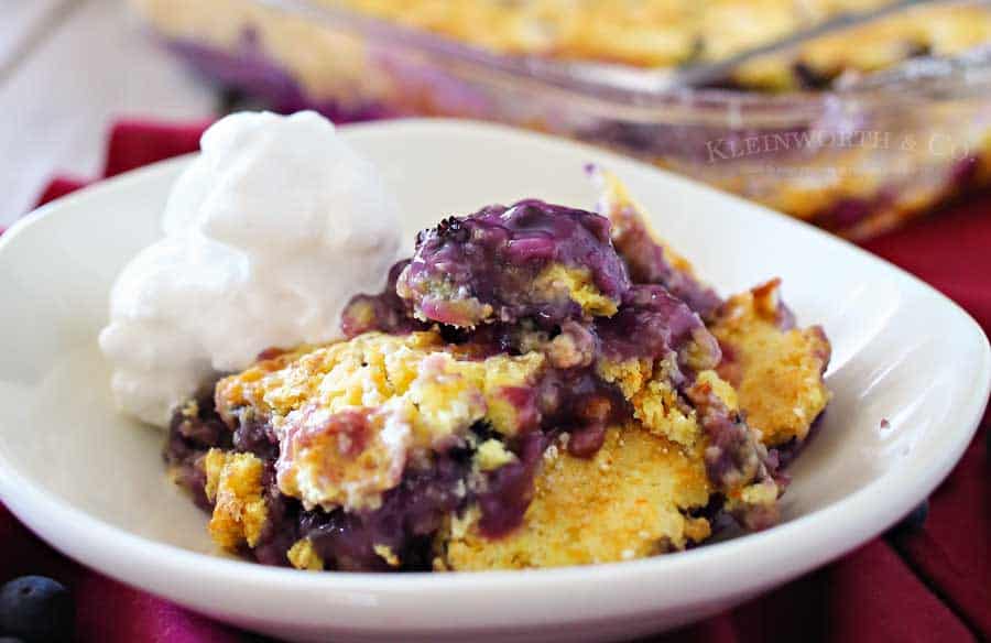 A serving of blueberry cobbler with a golden crust and a scoop of whipped cream on the side in a white bowl.