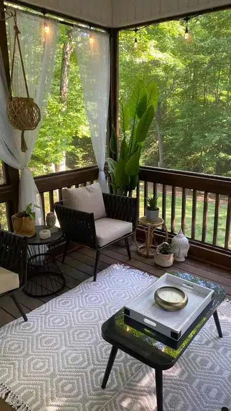 A screened-in porch features two black chairs with cushions, a small table, potted plants, a white rug, sheer curtains, and string lights, overlooking a green, wooded yard.