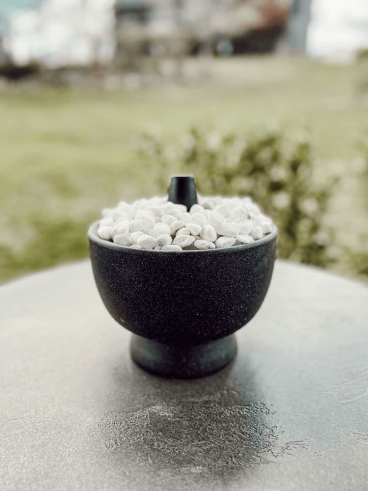 A black bowl filled with white pebbles sits on a round table outdoors, with blurred greenery in the background.
