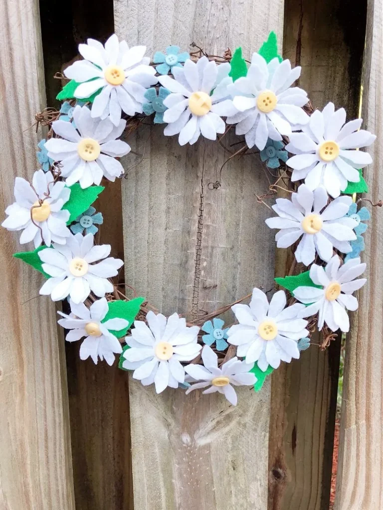 A wreath made of white felt flowers with yellow button centers and green felt leaves is hanging on a wooden fence.