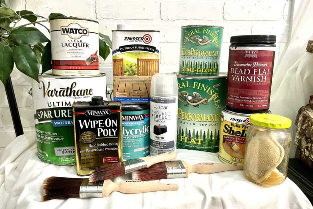 A variety of wood finishes, varnishes, sealers, spray paints, brushes, and applicators arranged on a white surface in front of a white brick wall.