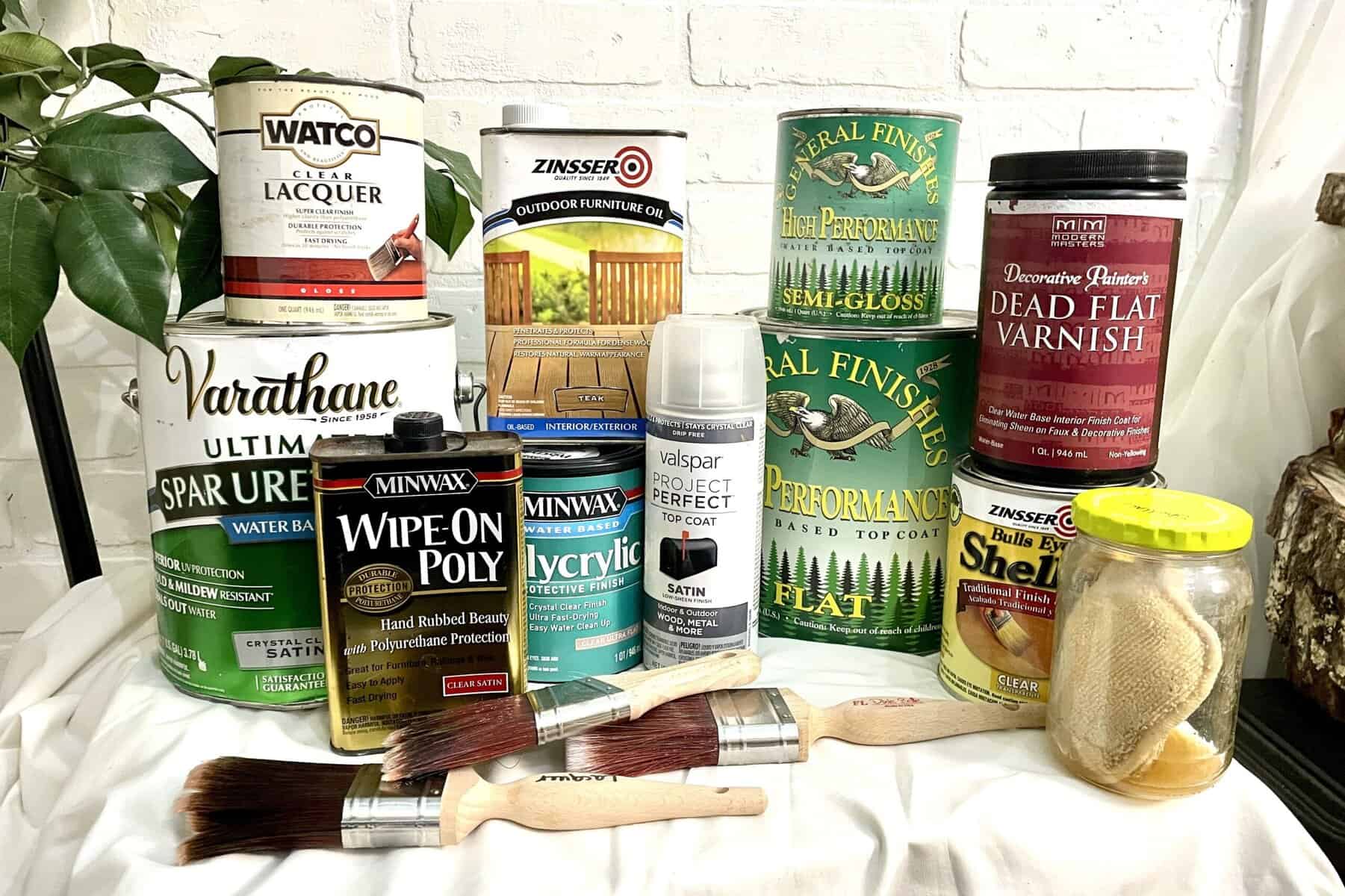 A variety of wood finishes, varnishes, sealers, spray paints, brushes, and applicators arranged on a white surface in front of a white brick wall.