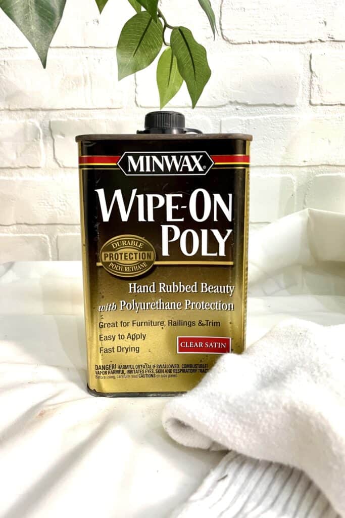 A can of Minwax Wipe-On Poly polyurethane protection rests on a white cloth with a leafy branch above, set against a white brick background.