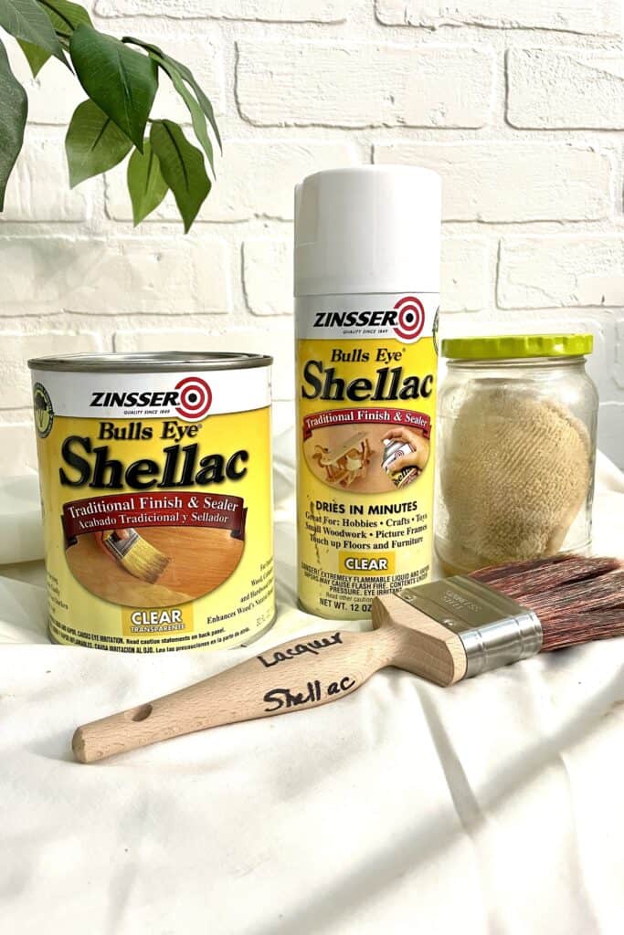 A can, spray can, and labeled brush of Zinsser Bulls Eye Shellac sit on a white cloth next to a glass jar with a cloth inside. A plant and white brick wall are in the background.