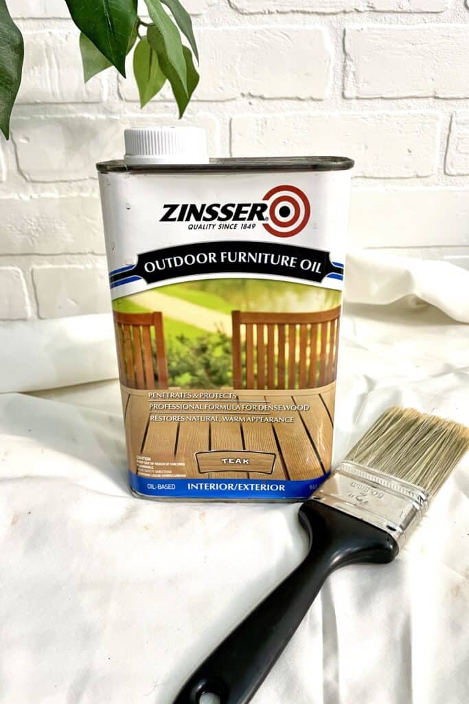 A can of Zinsser Outdoor Furniture Oil labeled "Teak" sits next to a paintbrush on a white surface, with a white brick wall and green leaves in the background.