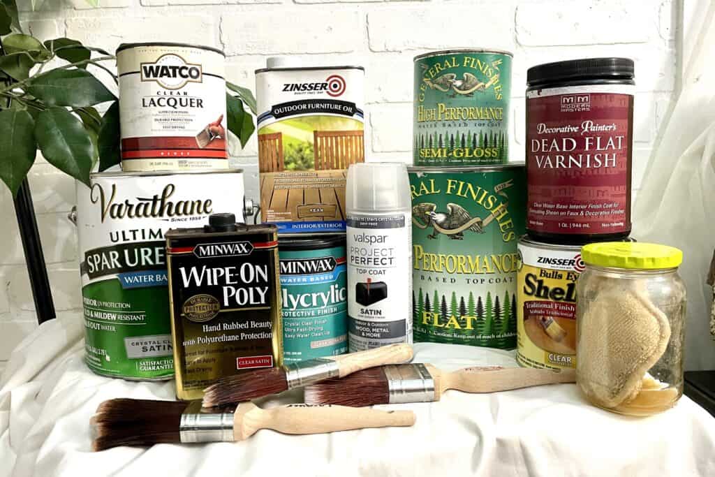 Assorted cans of wood finishes, varnishes, and sealants are displayed on a table with two paintbrushes, a spray can, and a jar of brushes or sponges in front of a white brick wall.