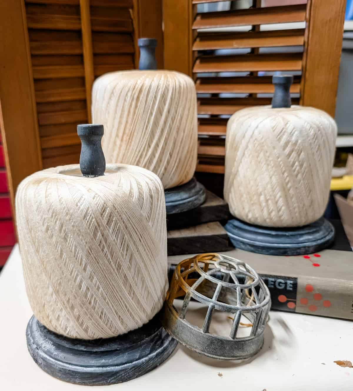 Three large spools of cream-colored string with dark holders are displayed on a table, alongside a metal cage-like object and a book. Wooden shutters are in the background.