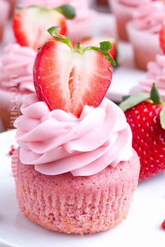 A pink cupcake with swirled strawberry frosting, topped with a halved strawberry, sits on a white plate with whole strawberries in the background.