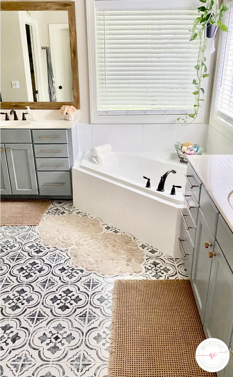 Bathroom with gray cabinets, patterned tile floor, a corner bathtub, two beige rugs, a vanity with a mirror, and a plant hanging near a window with blinds.
