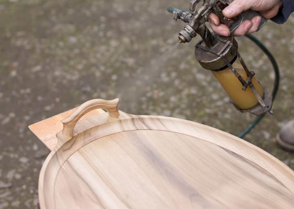A person uses a spray gun to apply finish to a wooden serving tray placed on an outdoor surface.