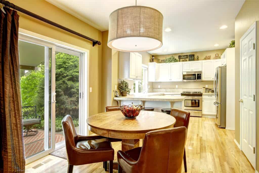 A modern kitchen and dining area with a round wooden table, four brown leather chairs, a large overhead light, and sliding glass doors opening to a patio and greenery outside.