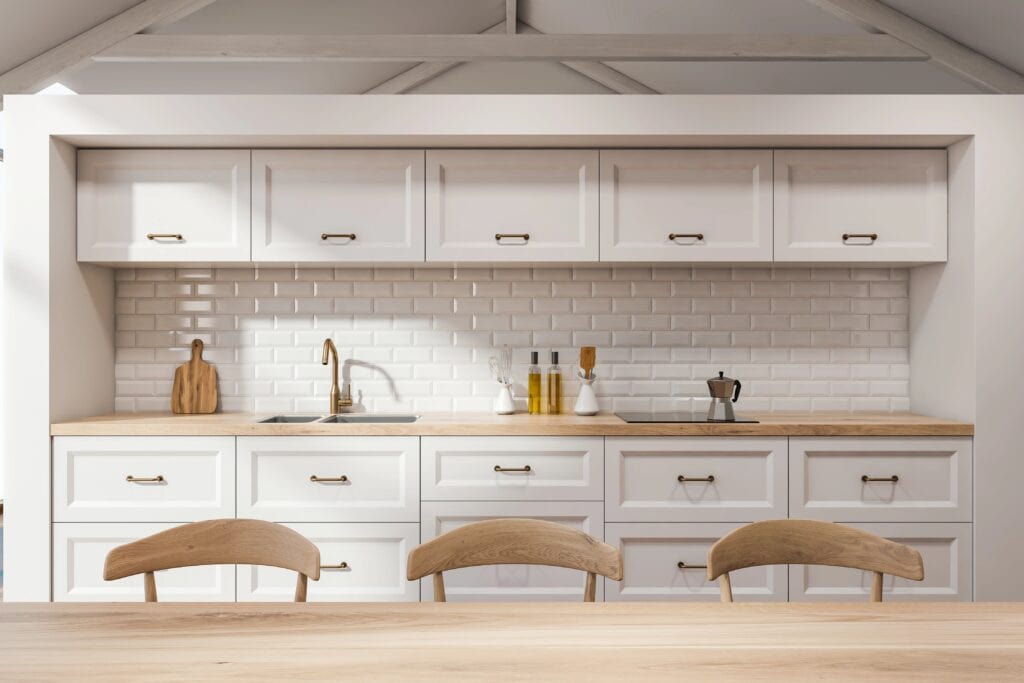 Modern white kitchen with wooden countertop, white cabinets, subway tile backsplash, sink, kitchenware, and three wooden chairs in the foreground.